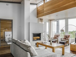 A modern living room with a sofa, coffee table, and chairs, a fireplace, and large windows.