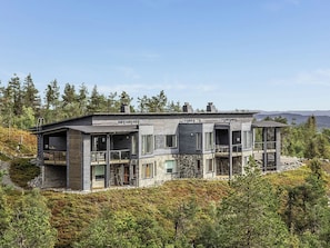 A modern two-story house with a stone facade, large windows, and a balcony overlooking a forested area.