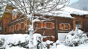 Una cabaña de madera con techo y ventanas cubiertas de nieve, rodeada de árboles y arbustos nevados.