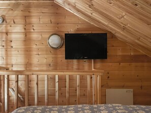 Ein Zimmer mit schrägverlaufender Holzbalkendecke, einem Wandfernseher, einem kleinen Tisch und einem Bett mit blumigem Überwurf.