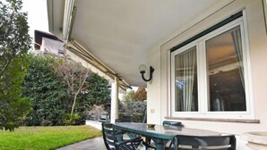 Una veranda esterna con tavolo e sedie, una finestra con tende e un giardino con diverse piante.