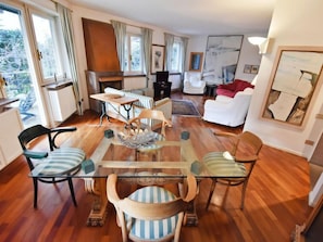 A spacious living area with a glass-top dining table surrounded by striped chairs, a fireplace, and a view of greenery outside. - Villa Esperia by Interhome (Merano)