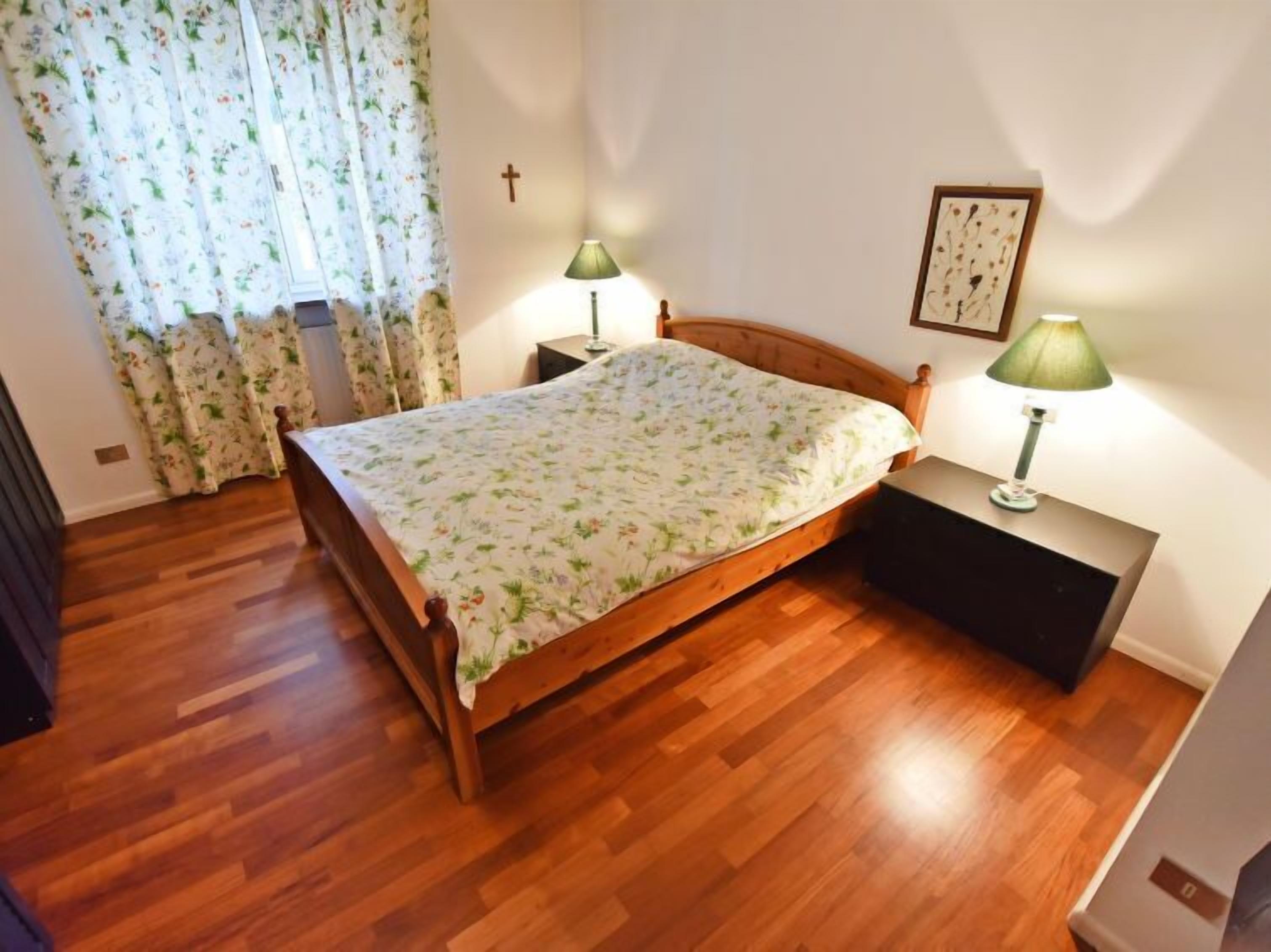 A bedroom with a wooden bed, floral bedspread, two bedside lamps, a cross on the wall, and a framed picture.