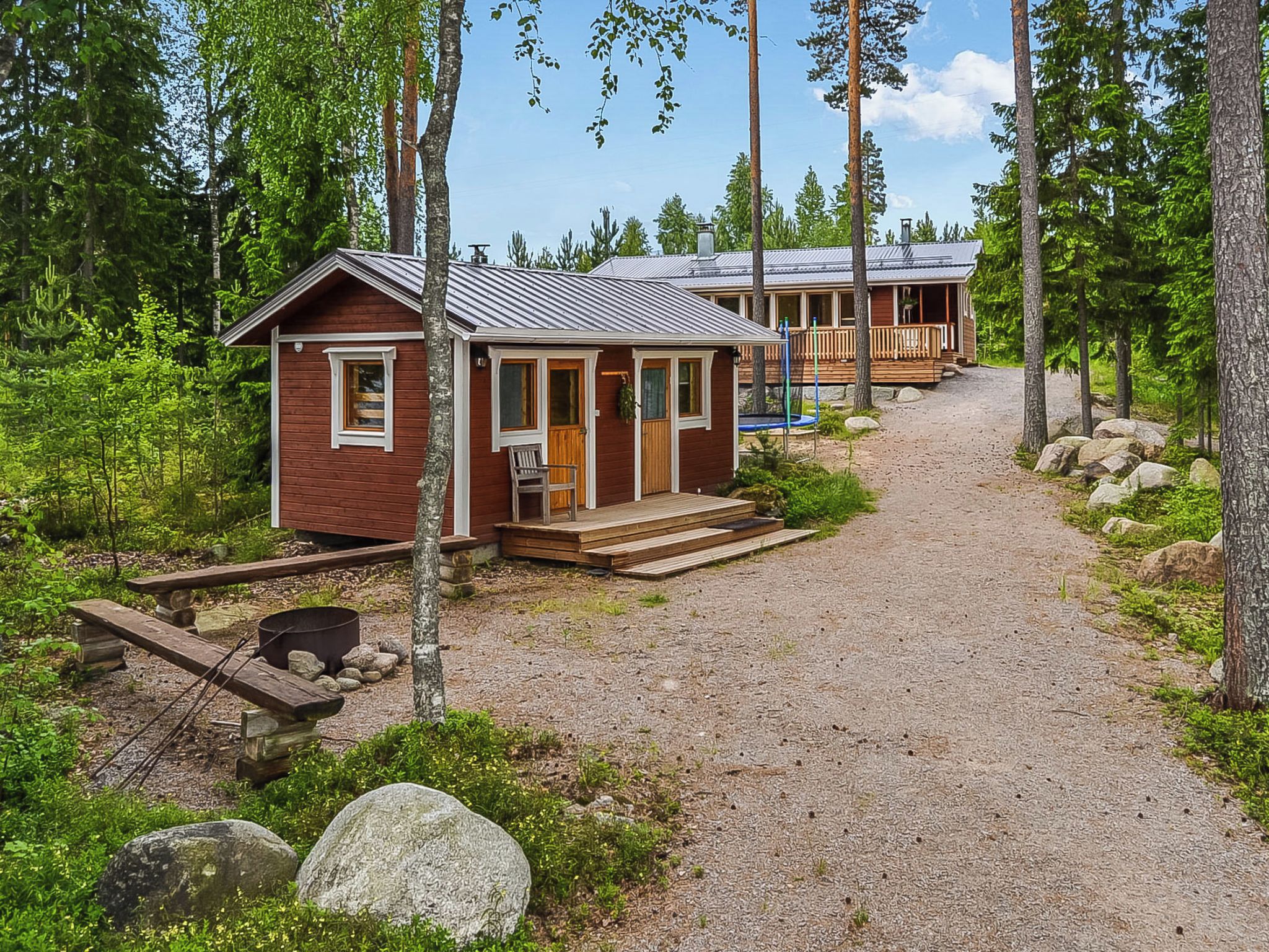 A cabin with a wooden deck, a gravel path leading to it, and a surrounding forest.