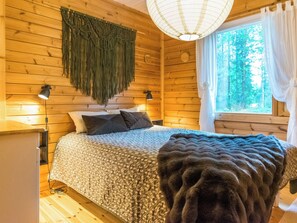 Een slaapkamer in een houten hutje, met een bed, twee bedlampjes, een macramé wanddecoratie, een raam met gordijnen en een plaid op de vloer.