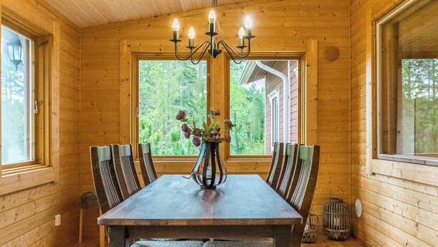 Interior de madera con una mesa de comedor, sillas, una lámpara de araña y ventanas con vistas a árboles.