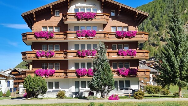 Ein mehrstöckiges Hotel mit Balkonen, die mit leuchtenden Blumen geschmückt sind, vor einer Kulisse aus üppiger Vegetation und blauem Himmel.