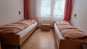Ein Zimmer mit zwei Einzelbetten, einem kleinen Nachttisch aus Holz, einem Fenster mit durchsichtigen Vorhängen und Blick auf Gebäude.