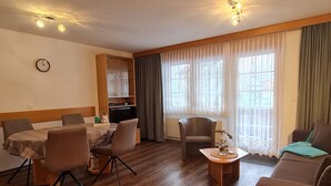 Ein Wohnzimmer mit Essbereich, Holzboden, einer Couch, einem Sessel, einem kleinen Tisch mit einer Vase und einer Uhr an der Wand.