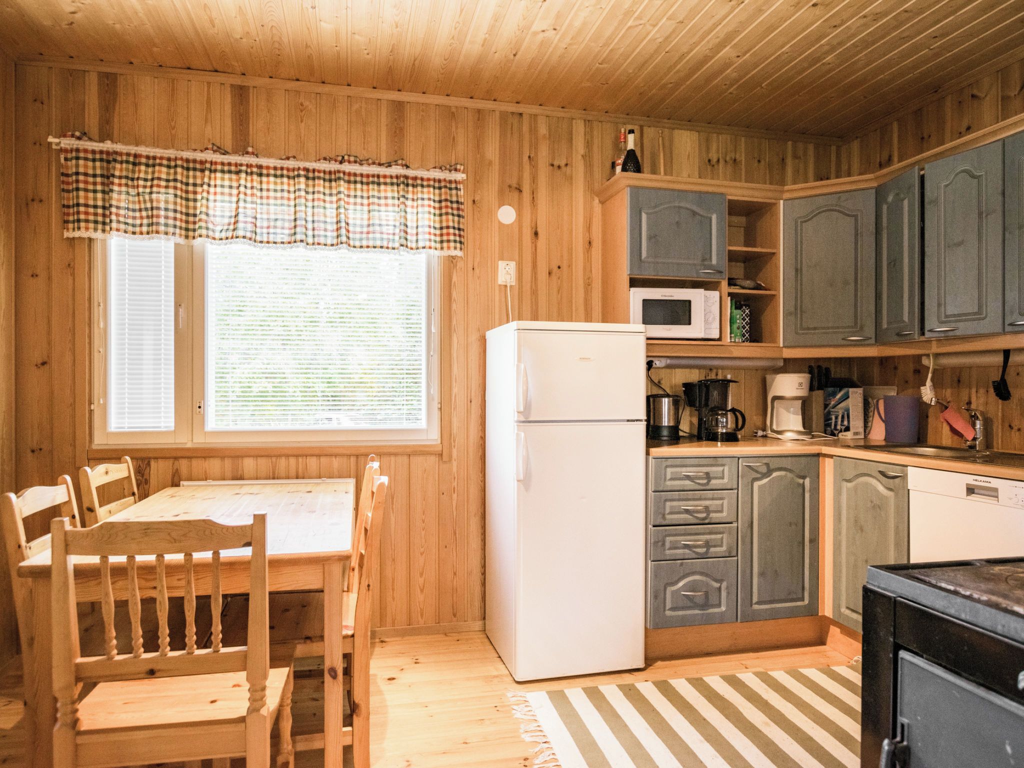 A wooden-paneled kitchen with a white refrigerator, a microwave, and a coffee maker. There is a dining table with chairs and a window with blinds.