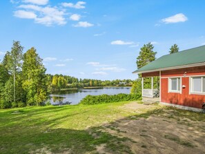 Una cabana al costat d'un llac amb un porxo, envoltada d'arbres i sota un cel clar.