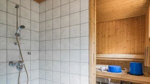 Un espace douche équipé d’un pommeau de douche à main et d’un robinet mural. On y trouve une étagère en bois sur laquelle sont posés un récipient bleu et un objet blanc.