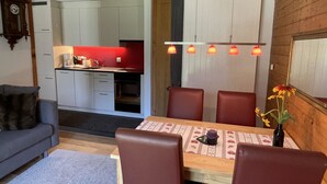 A dining area with a wooden table, four chairs, and a vase with flowers. A kitchen is visible in the background with white cabinets and a red countertop. - Sweda EG Süd by Interhome (Fiesch)