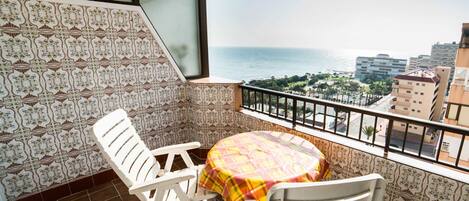 A balcony with a view of the sea and cityscape, featuring patterned tiles, white chairs, and a round table with a colorful tablecloth.