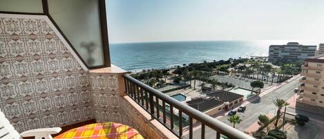 A balcony with a patterned tile wall, a view of the sea, and a parking area with palm trees.