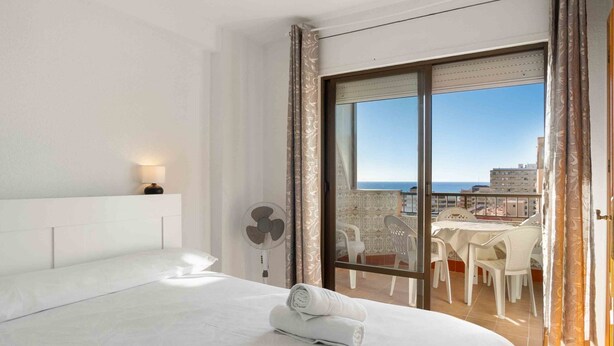 Ein Hotelzimmer mit Bett, Ventilator und Blick auf Stadt und Meer durch ein großes Fenster.