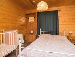 A wooden cabin bedroom with a bed, a crib, a nightstand, a lamp, and a window with curtains. - Iltaniemi by Interhome (Enonkoski)