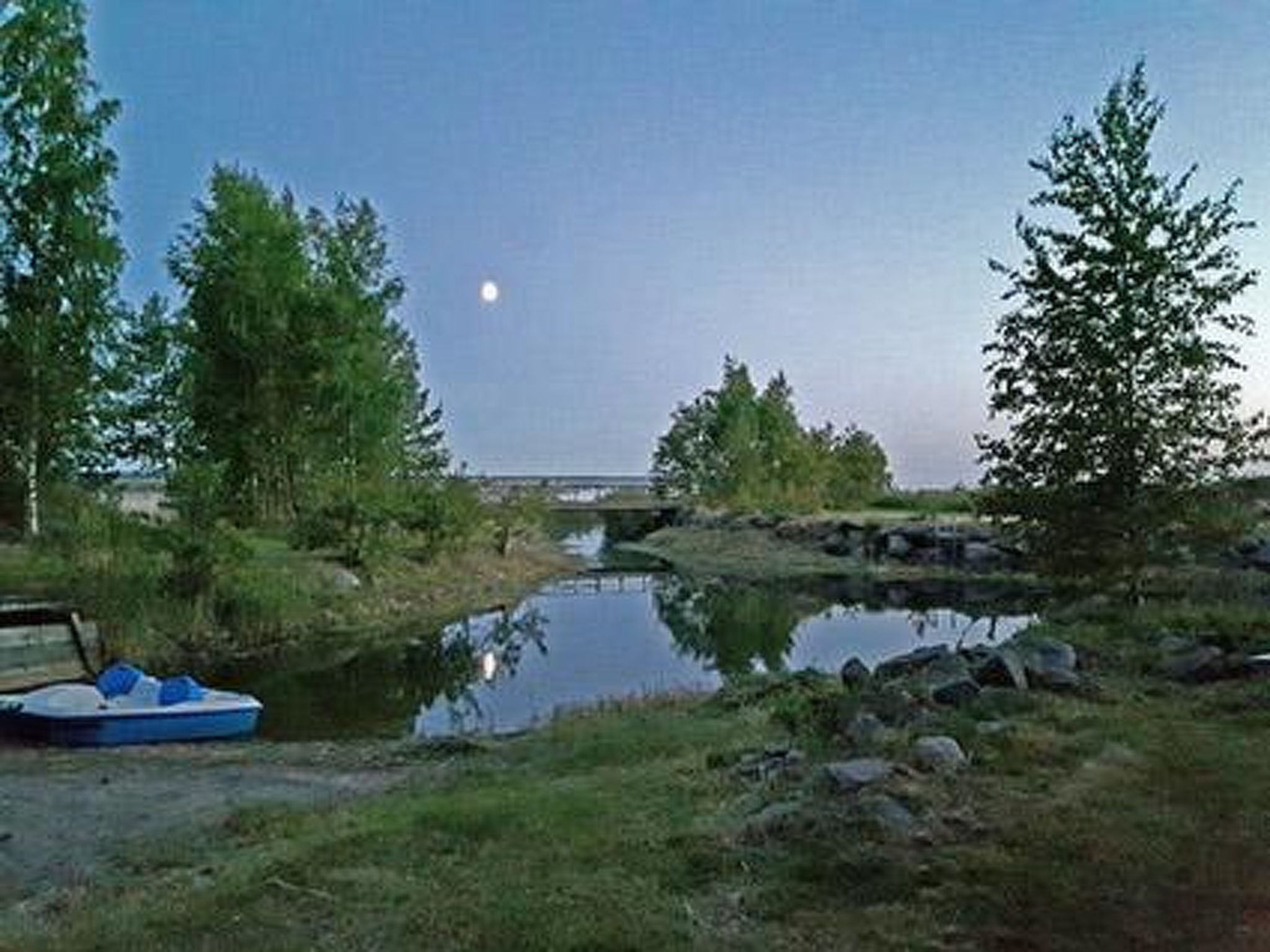 Cena serena à beira de um lago, com um barco atracado, rodeado por árvores e uma pequena ponte ao fundo.