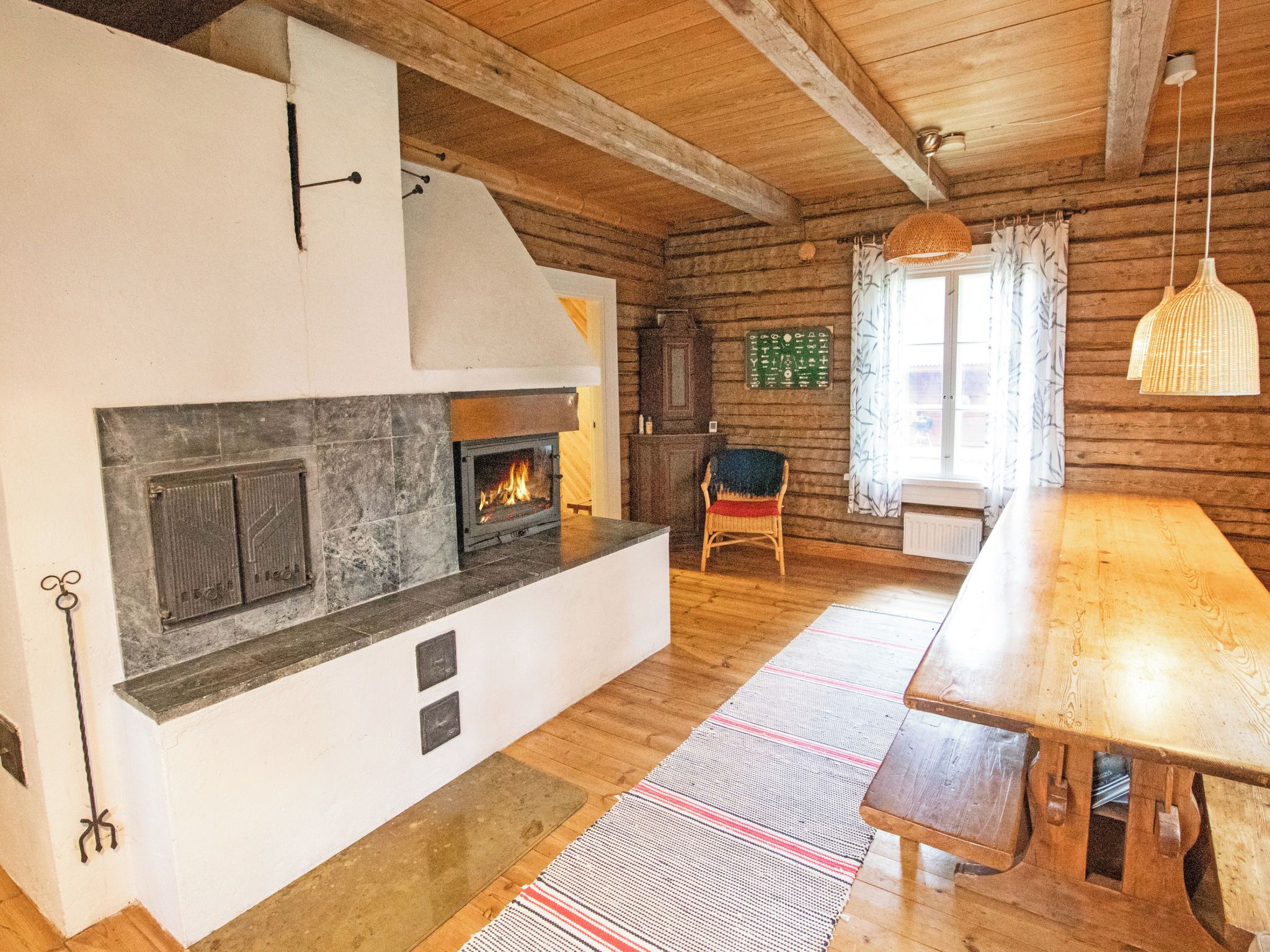 A cozy room with a fireplace, wooden beams, a dining table, and a window with curtains.
