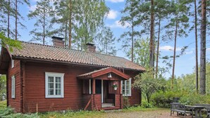 Une cabane en bois avec un toit en tuiles, entourée de grands arbres et bordée d’un chemin de gravier.