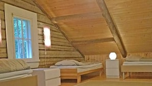 Ein gemütliches Schlafzimmer im Dachgeschoss mit schrägen Wänden, Holzboden und einem Fenster mit Blick auf Bäume. Es gibt zwei Betten, einen Nachttisch und eine Lampe.