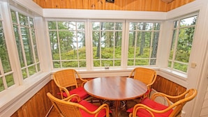 Une pièce d’angle avec une table ronde et six chaises en osier dotées de coussins rouges, de grandes fenêtres offrant une vue sur les arbres et des murs en bois.