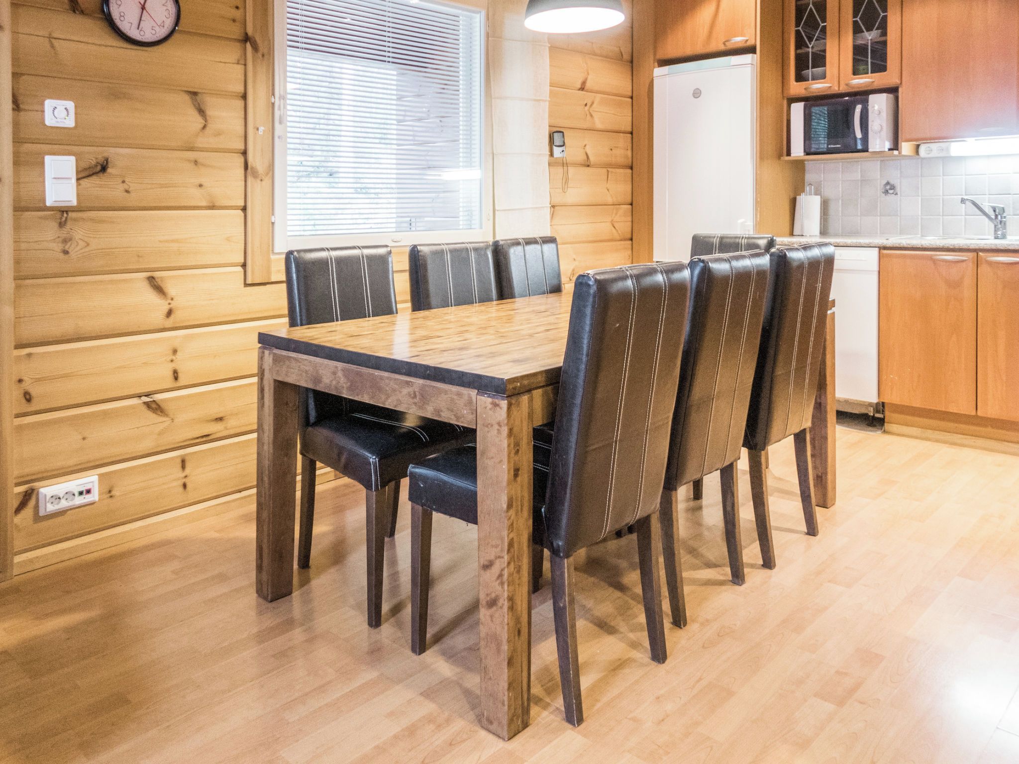 Habitación con paneles de madera, una mesa de comedor con sillas, un reloj y una zona de cocina con electrodomésticos.