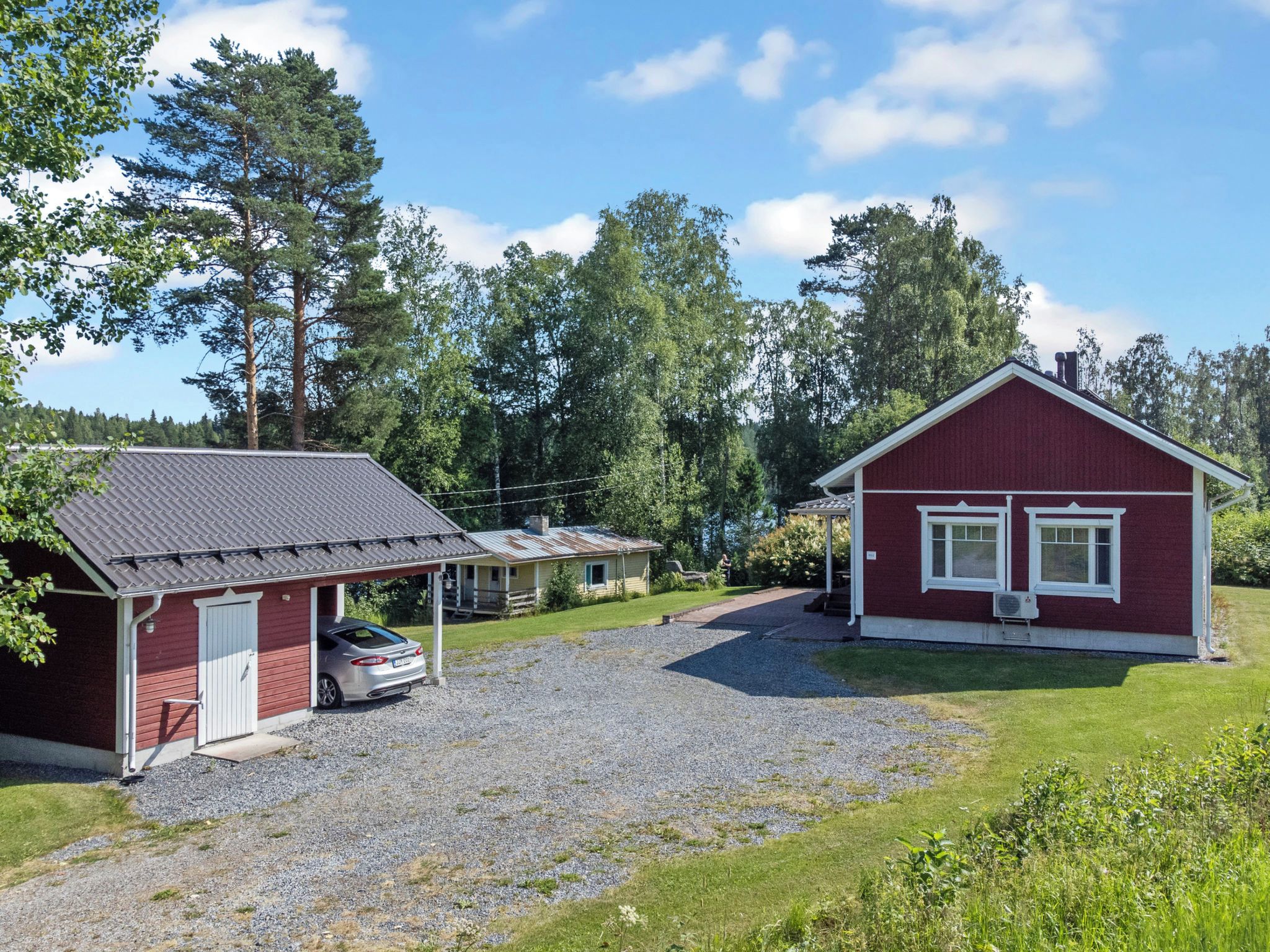 A red building with a car parked in front, surrounded by trees and a gravel driveway.