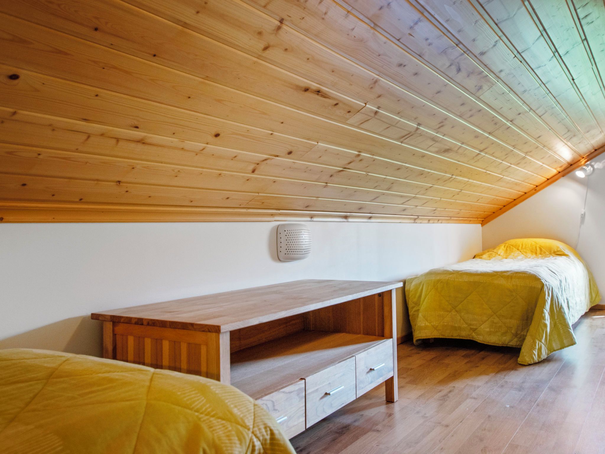 Une chambre avec un plafond en pente, un plancher en bois, un lit avec une literie jaune, une table de chevet et un haut-parleur fixé au mur.