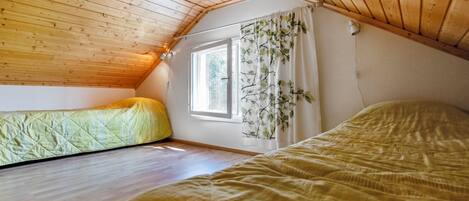 Ett litet, mysigt sovrum på vinden med träribbor i taket, ett fönster med gardiner och två sängar.