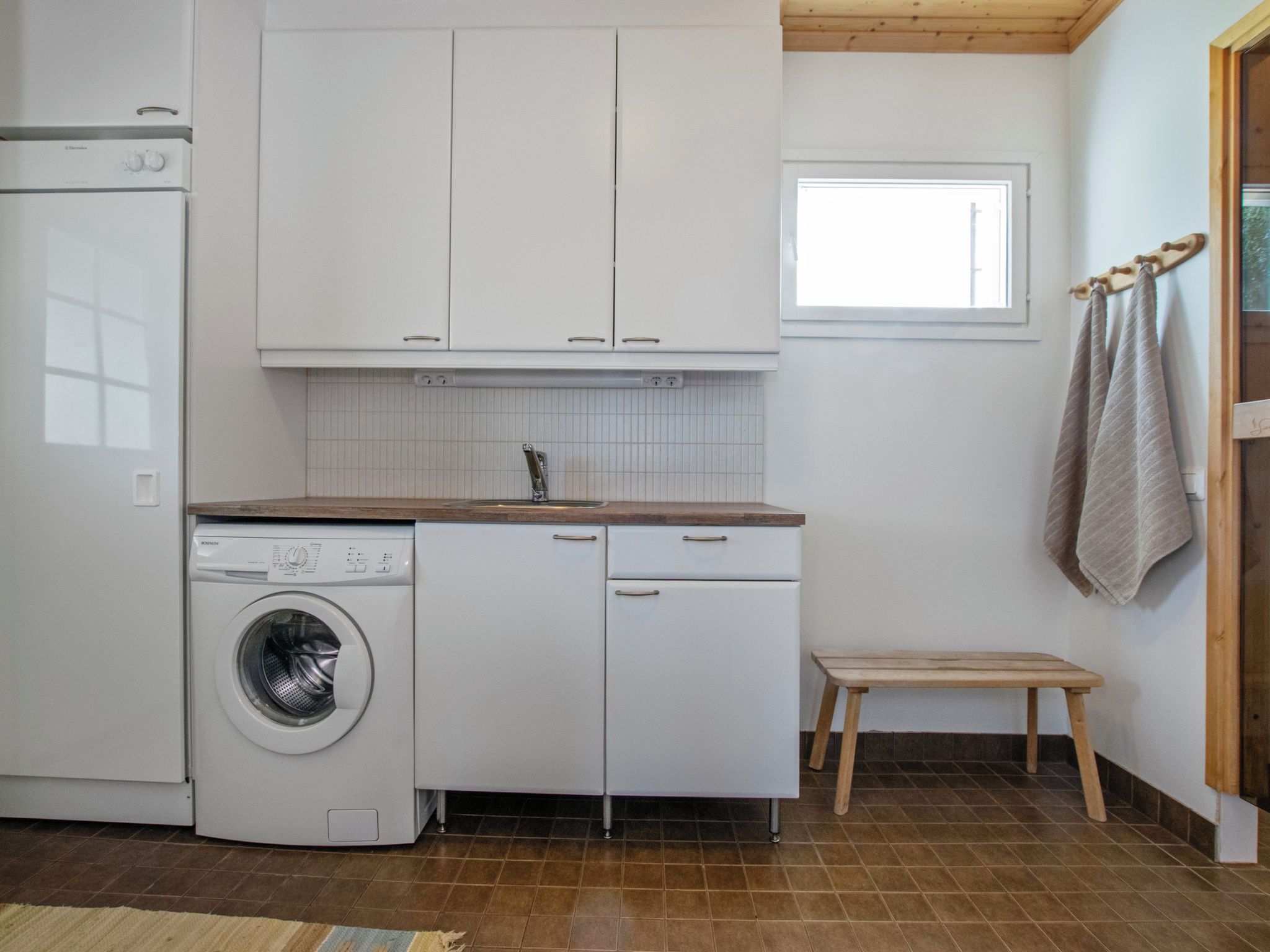 A compact kitchen with a washing machine, a sink, and a small wooden stool.