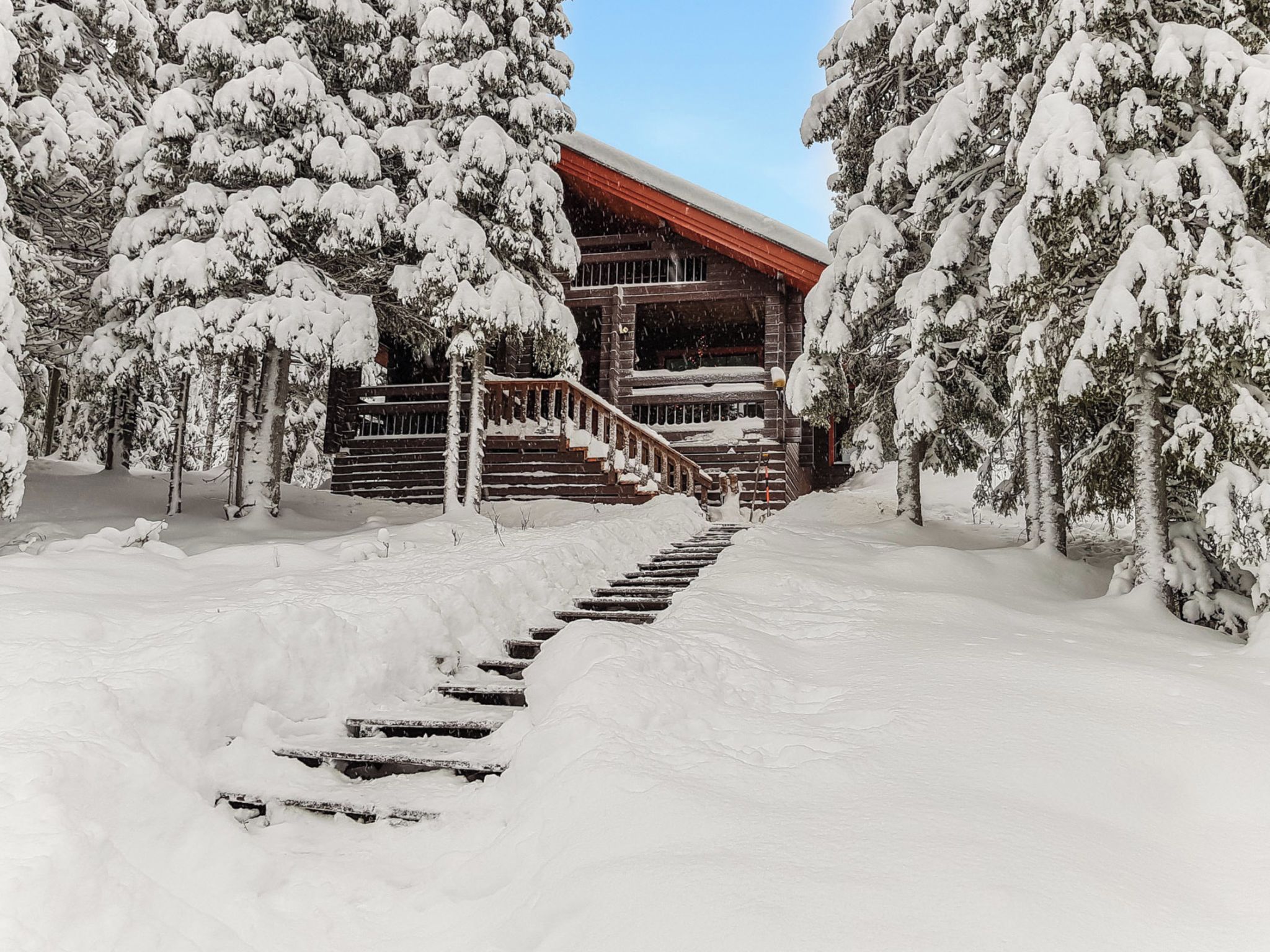 Eine schneebedeckte Holztreppe führt zu einer Hütte mit rotem Dach, umgeben von schneebedeckten Bäumen.