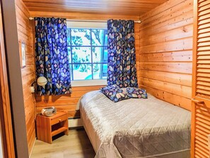 A small wooden room with a bed, a nightstand, a window with curtains, and a view of greenery outside.