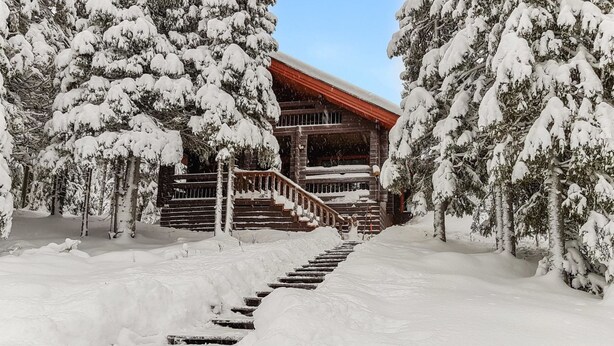 A snow-covered wooden cabin with a staircase leading to a red-roofed entrance.