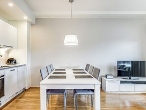 A modern kitchen with white cabinets, a dining table with chairs, a hanging light fixture, and a television on a stand.