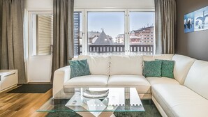 Un salon avec un canapé beige, une table basse en verre et une vue sur les immeubles à travers de grandes fenêtres.