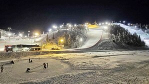 A snow-covered ski slope at night with artificial lighting, a ski lift, and a few people.