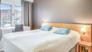 Une chambre d’hôtel avec un grand lit, un bureau et une chaise. Il y a une fenêtre avec des rideaux et une lampe fixée au mur.
