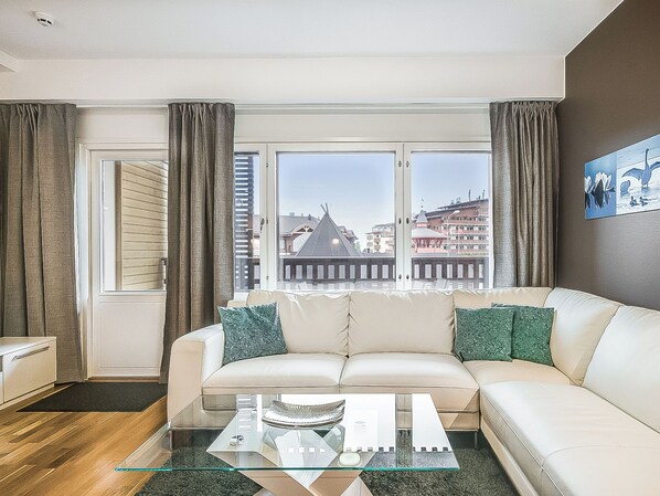 A modern living room with a sofa, a glass coffee table, and a view of buildings through large windows.