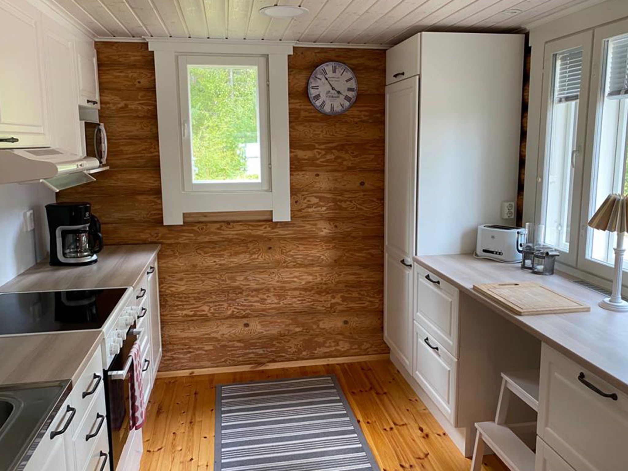 A compact kitchen with wooden walls, a built-in stove, a coffee maker, and a clock on the wall.