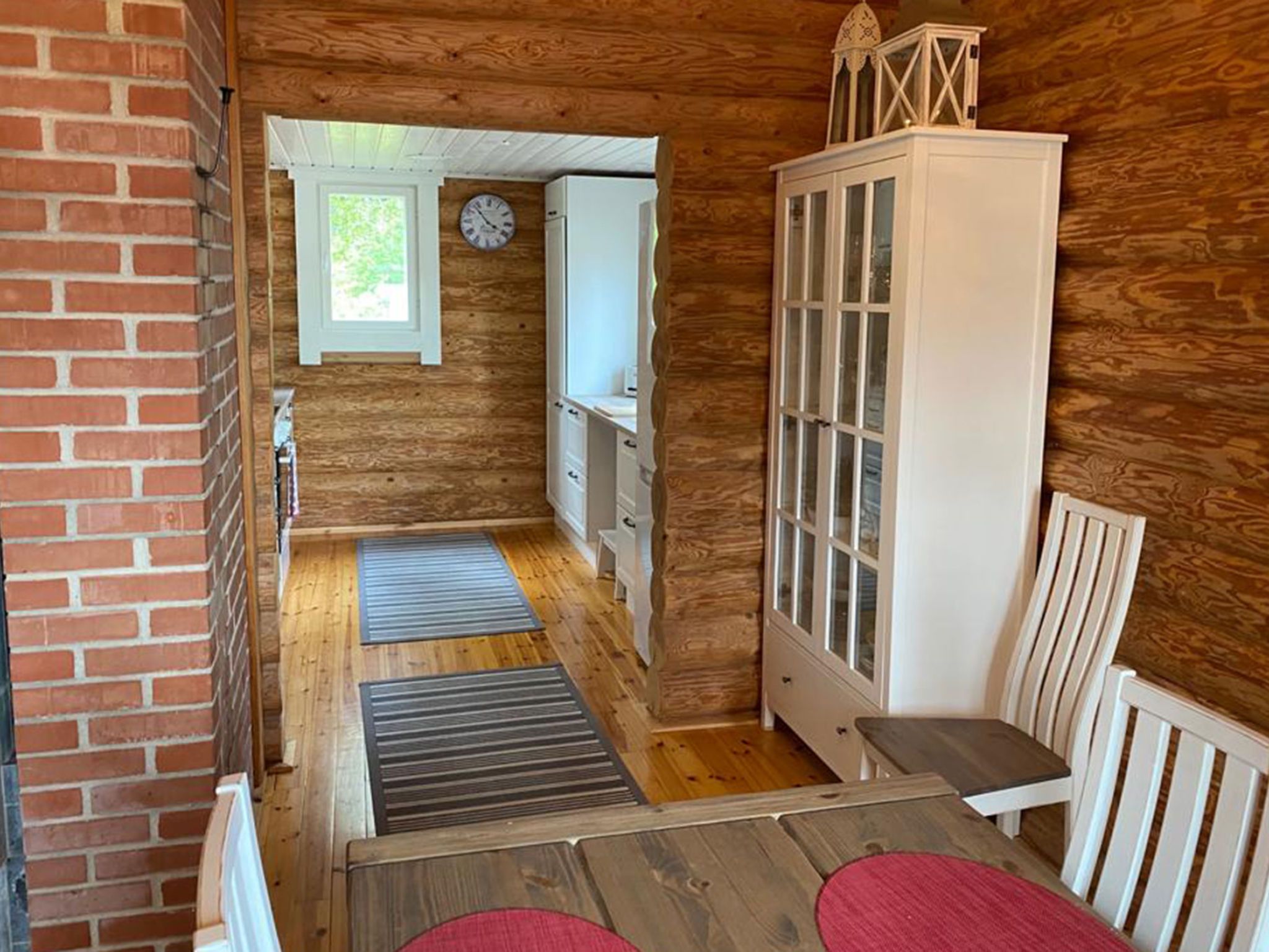 A compact wooden room with a dining table, a cabinet, a clock, and a window.