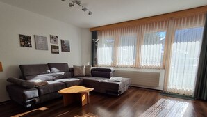 A modern living room with a sectional sofa, a wooden coffee table, and a large window with sheer curtains. - Apartment Orion by Interhome (Saas-Grund)