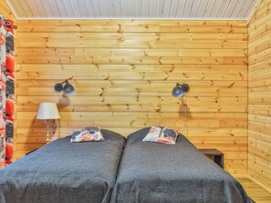 Huone, jonka seinät ovat puuta, ja jossa on kaksi harmaaliinavaatteilla varustettua sänkyä, lamppu ja pieni pöytä.