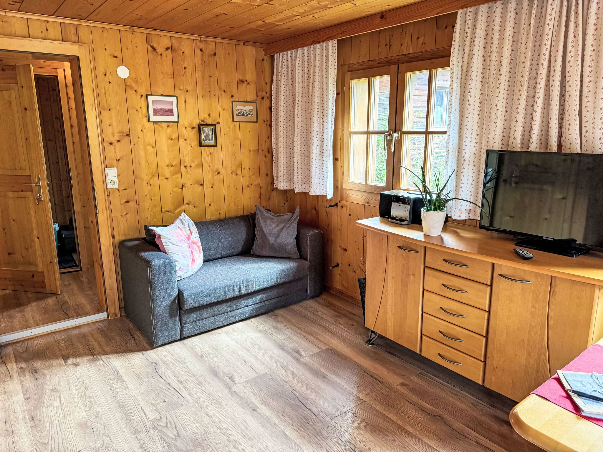 L’intérieur d’un chalet accueillant, avec un plancher en bois, un canapé gris agrémenté de coussins, un meuble en bois doté de tiroirs, un téléviseur et une fenêtre ornée de rideaux.