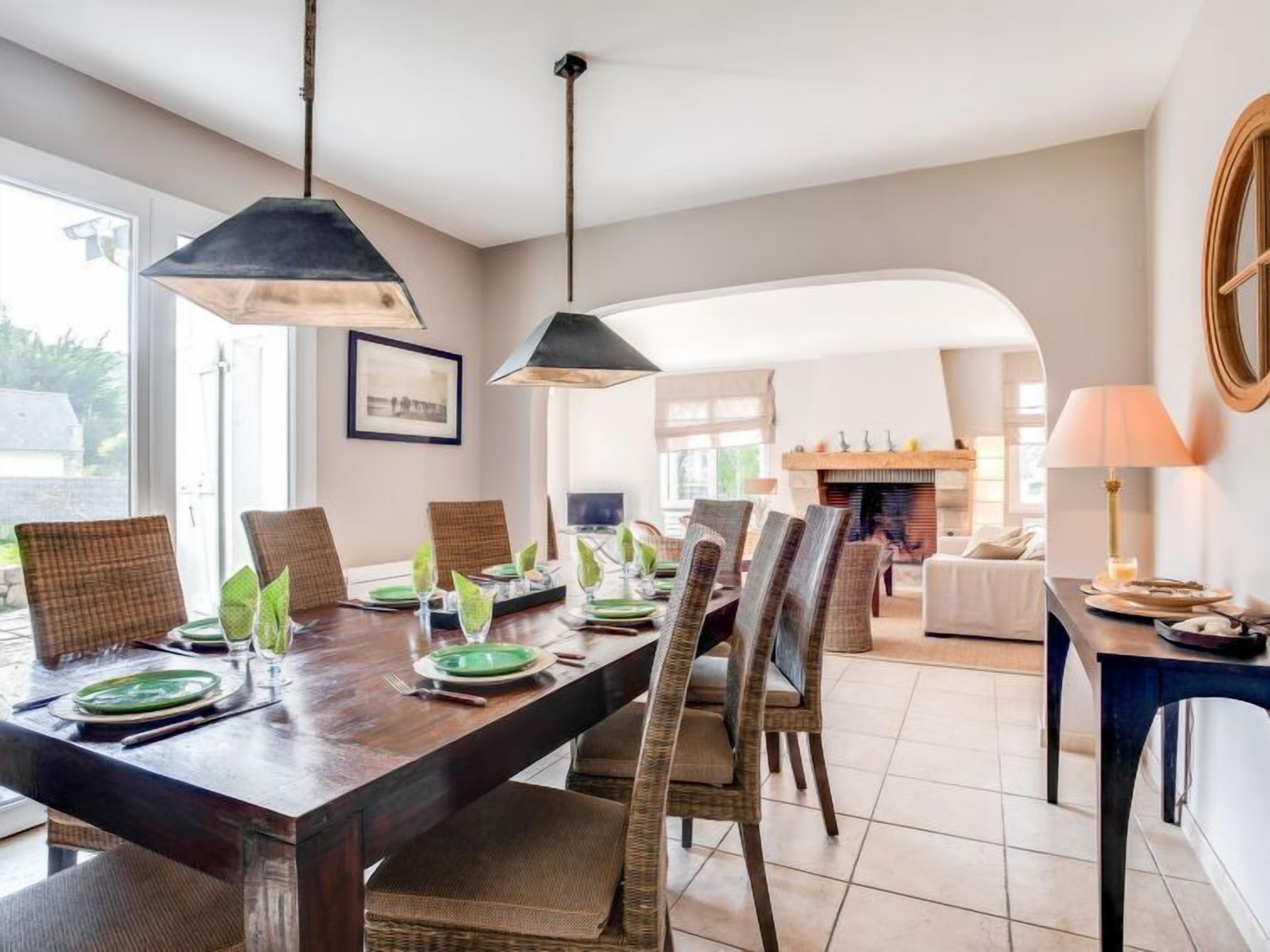 A dining area with a wooden table set for four, surrounded by wicker chairs. There is a fireplace in the background, and the room has a modern, clean design with a large window providing natural light.
