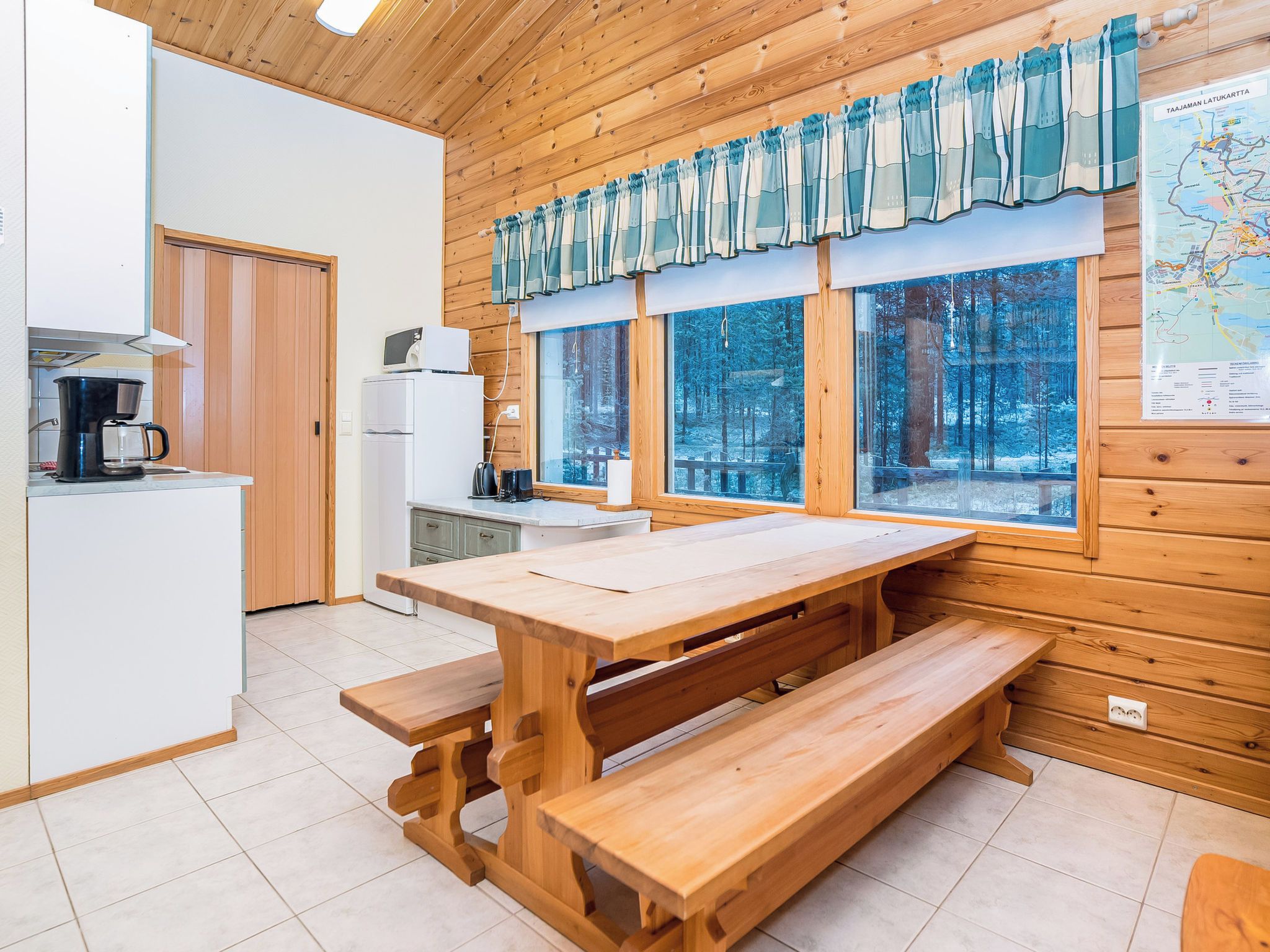 A wooden interior with a kitchen area, a dining table with benches, and large windows overlooking a forest.