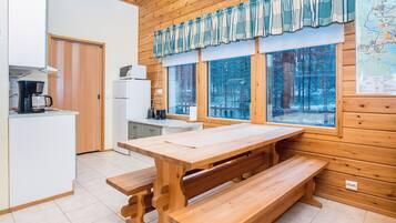 A wooden interior with a kitchen area, a dining table with benches, and large windows overlooking a forest.