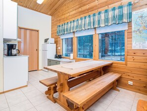 Interior de fusta amb zona de cuina, taula de dinar amb bancs i grans finestres amb vista a un bosc.