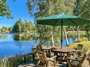 A wooden deck with a table and chairs, an umbrella, and a view of a lake with trees and a house in the background. - Hartolan ilo by Interhome (Hartola)