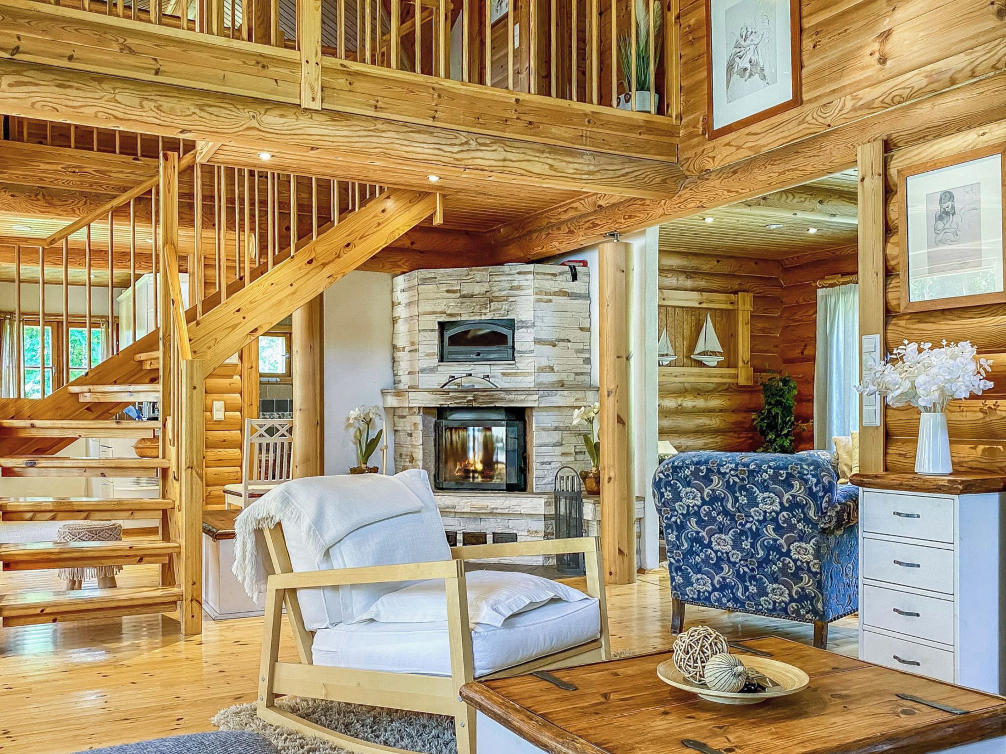 A log cabin interior with a stone fireplace, wooden staircase, and a cozy seating area.